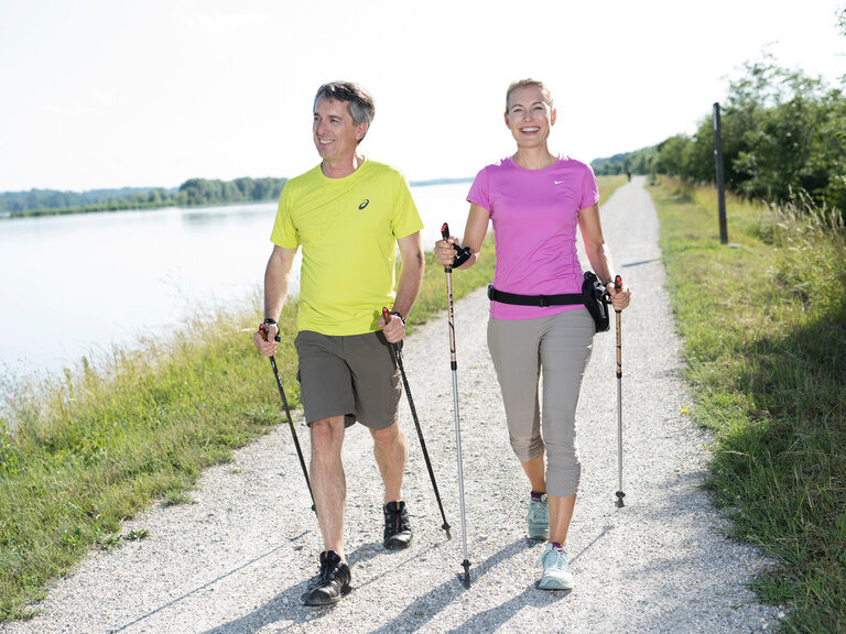 Ein Mann und eine Frau spazieren gemeinsam am Flussufer des Inns mit Stöcken entlang.